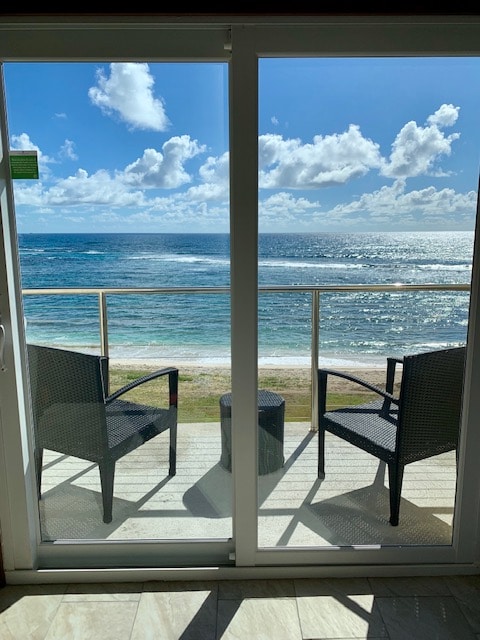 Two outdoor chairs are positioned on a private terrace, which overlooks the expansive ocean. A small table sits between the chairs, framing the view of the water and sky filled with scattered clouds. The scene offers a tranquil atmosphere with a clear blue horizon.