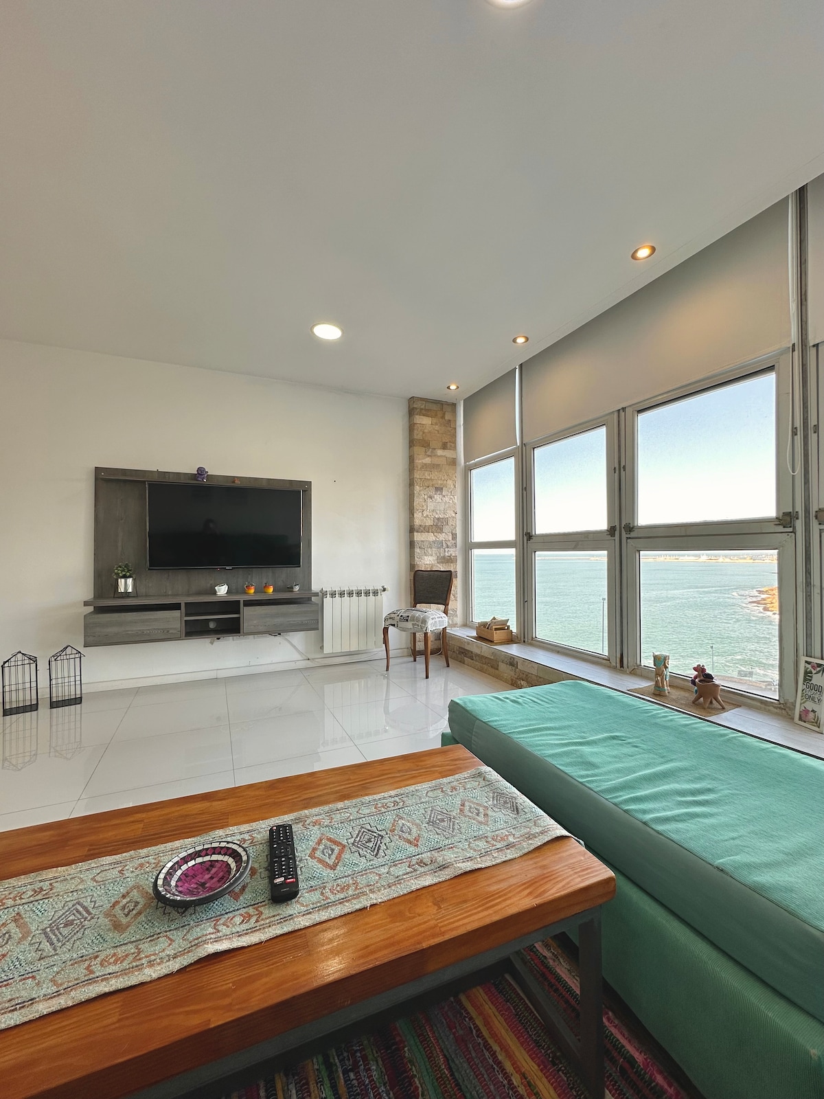 The living area features large windows offering a view of the water and ample natural light. A comfortable sofa is positioned next to a wooden table, with a TV mounted on the wall. Decorative elements are placed nearby, enhancing the space.