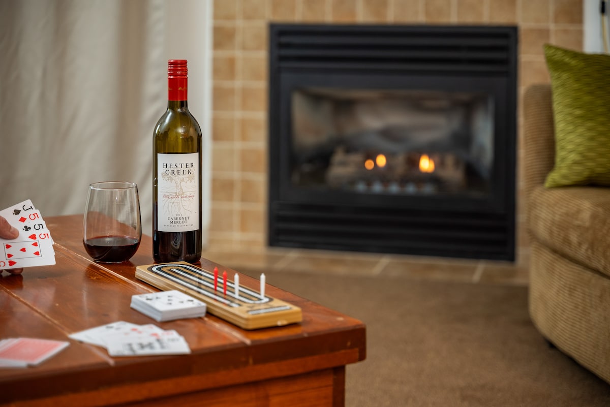 A warm indoor scene features a cozy fireplace with a soft glow. A bottle of wine and a glass sit on a wooden table alongside a card game, inviting relaxation and leisure. The rich texture of a green accent pillow complements a tan sofa.