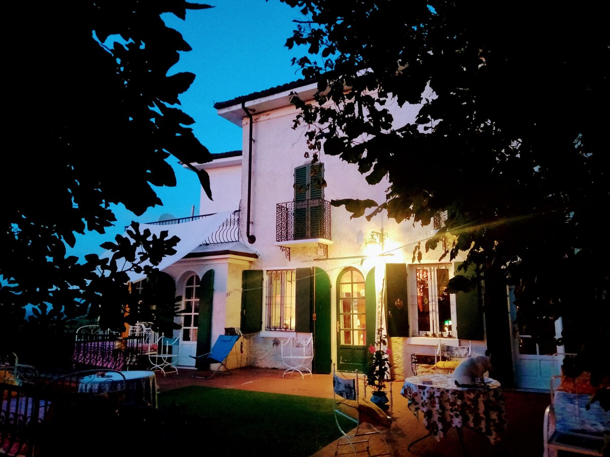 The villa is softly illuminated in the evening light, showcasing its charming exterior with green shutters and multiple windows. Outdoor furniture is arranged on the terrace, surrounded by lush greenery, creating a serene setting for relaxation.