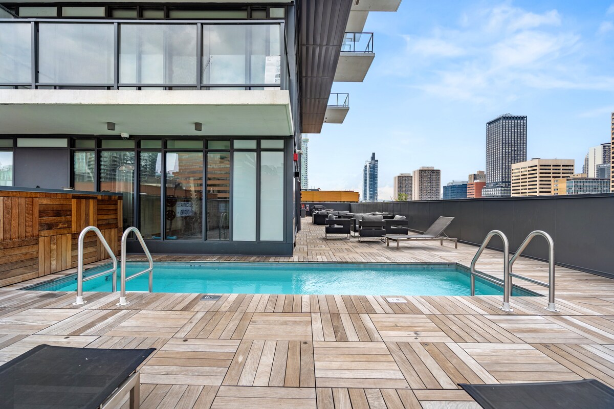 An outdoor rooftop pool is framed by wooden decking and sleek lounge furniture. The skyline of downtown Calgary, including high-rise buildings, is visible in the background. Glass railings protect the area while providing unobstructed views of the city.