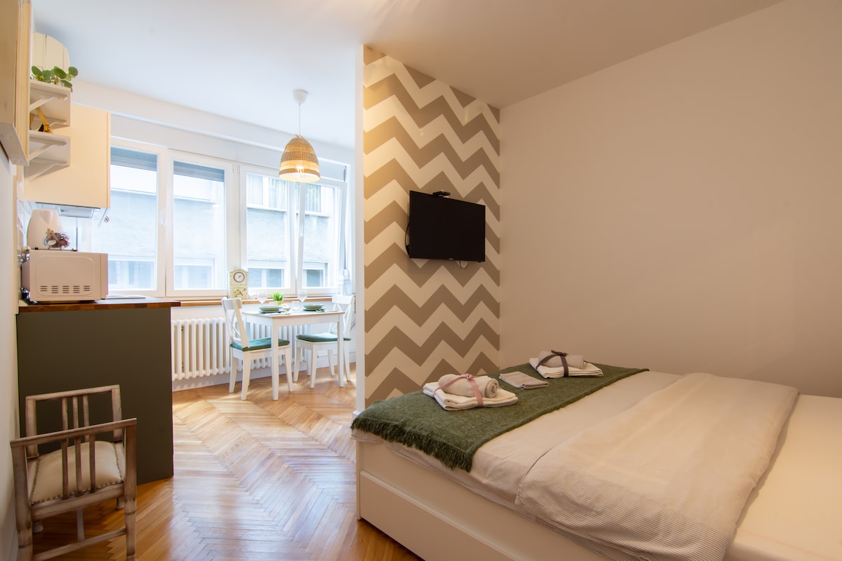 A bright sleeping area features a spacious bed covered with a light blanket. A television is mounted on a patterned wall, while a dining table and chairs are visible by the window, allowing natural light to fill the room.