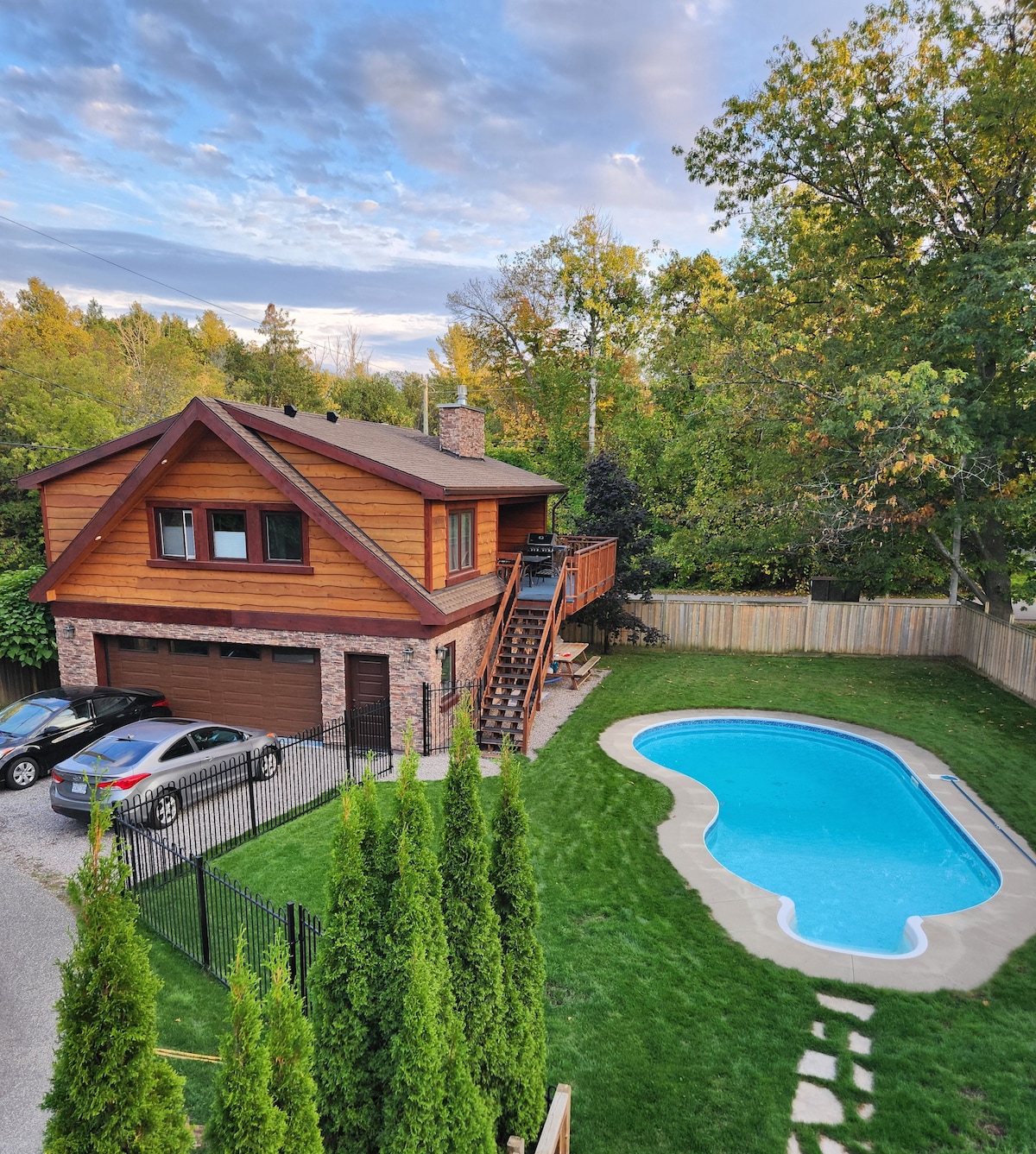 An aerial view of the rustic chalet reveals a warm wooden exterior complemented by brick accents. A fenced-in yard features a heated, welcoming saltwater pool. Two cars are parked in the driveway, and greenery from surrounding trees enhances the serene atmosphere.