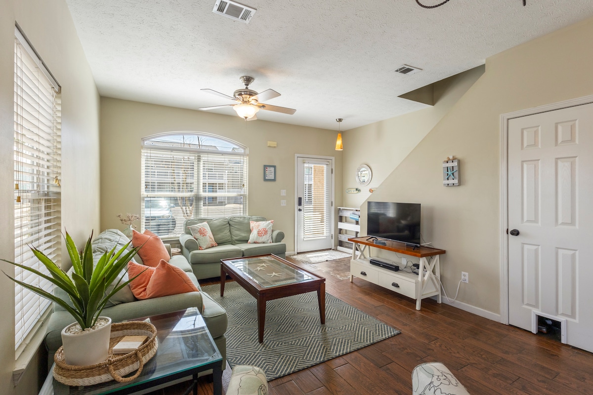 The inviting living area features a comfortable seating arrangement with a light green sectional sofa and a wooden coffee table. A flat-screen TV is situated on a white media console. Natural light fills the space through large windows with blinds, enhancing the relaxed atmosphere.