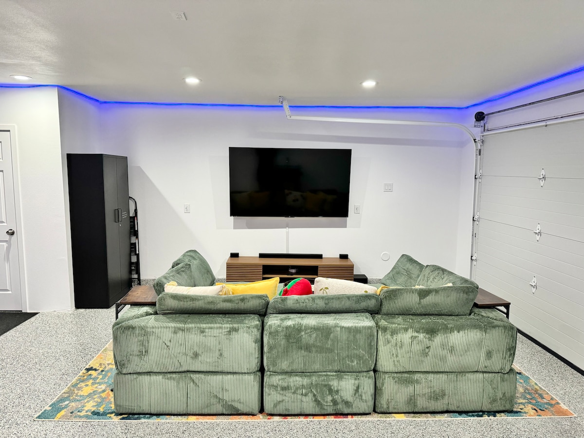 A game room is shown with a large sectional sofa upholstered in green fabric facing a wall-mounted TV. Blue LED lighting is installed around the ceiling's perimeter, and a textured area rug adds color to the space. A closet is visible to the side.