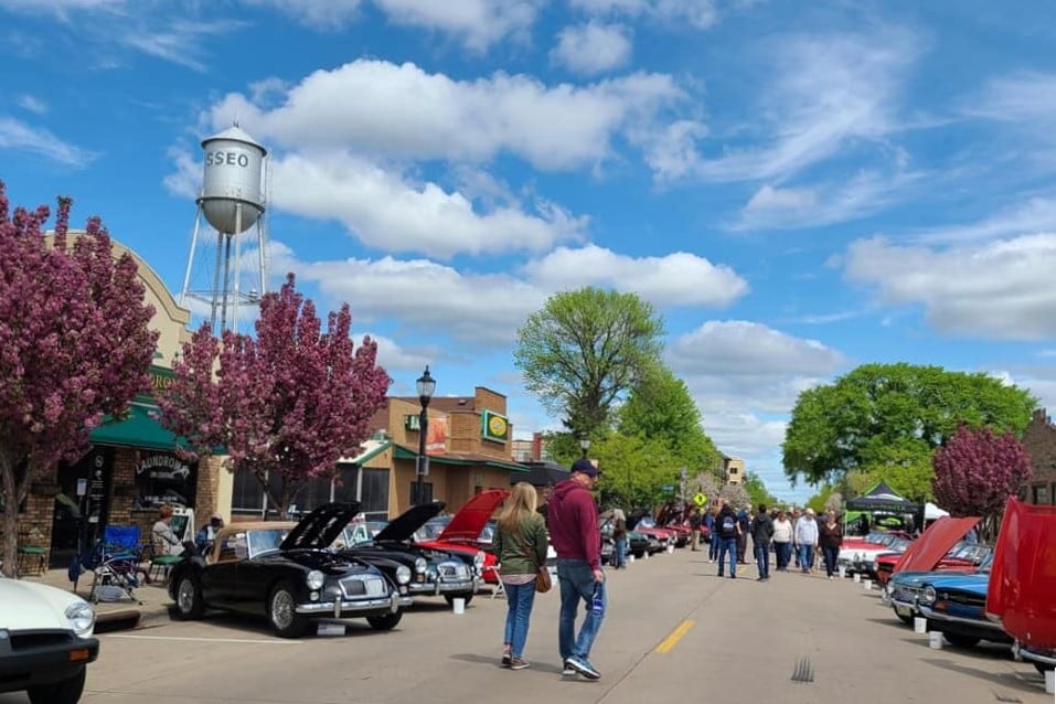 A vibrant street scene in Downtown Osseo features classic cars lined along the road. Visitors stroll among blooming trees and historic buildings under a clear blue sky, with fluffy white clouds adding depth to the atmosphere.