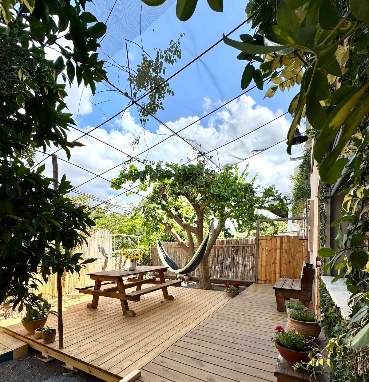 An outdoor space features a wooden deck shaded by a canopy. A picnic table is positioned at the center, with a hammock hanging from a tree nearby. Potted plants are arranged around the area, providing a green and tranquil setting.