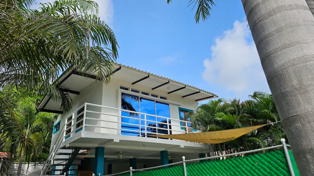 A two-story house is presented with large windows reflecting the sky. The structure is surrounded by lush green palm trees, and a fabric awning stretches across the outdoor space below. The inviting exterior features clean lines and a modern design.