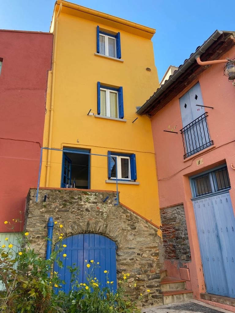 A three-story house features a vibrant yellow facade complemented by blue window frames and a blue wooden door. An exterior stone staircase leads to the entrance, highlighting the charming architecture of the surrounding pastel-colored buildings.