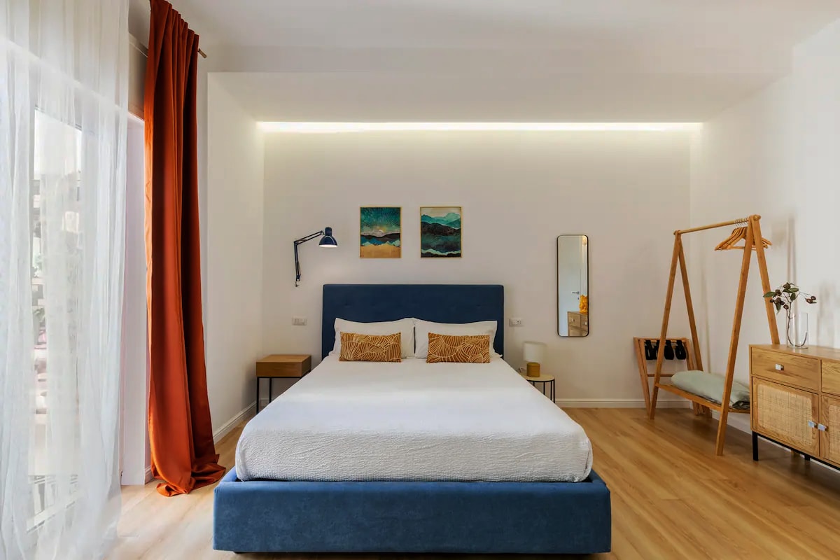 A spacious bedroom is featured with a modern blue bed frame and soft, white bedding. Natural light flows through large windows draped with light curtains, illuminating artworks on the wall. A wooden wardrobe and a bedside table provide functional decor, enhancing the room's serene atmosphere.