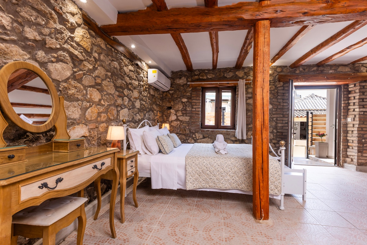 The cozy studio features a spacious bed dressed in neutral tones, complemented by a wooden headboard and decorative pillows. Stone walls and wooden beams create a rustic charm. A vintage dresser and mirror add elegance, while light filters through the window, brightening the room.