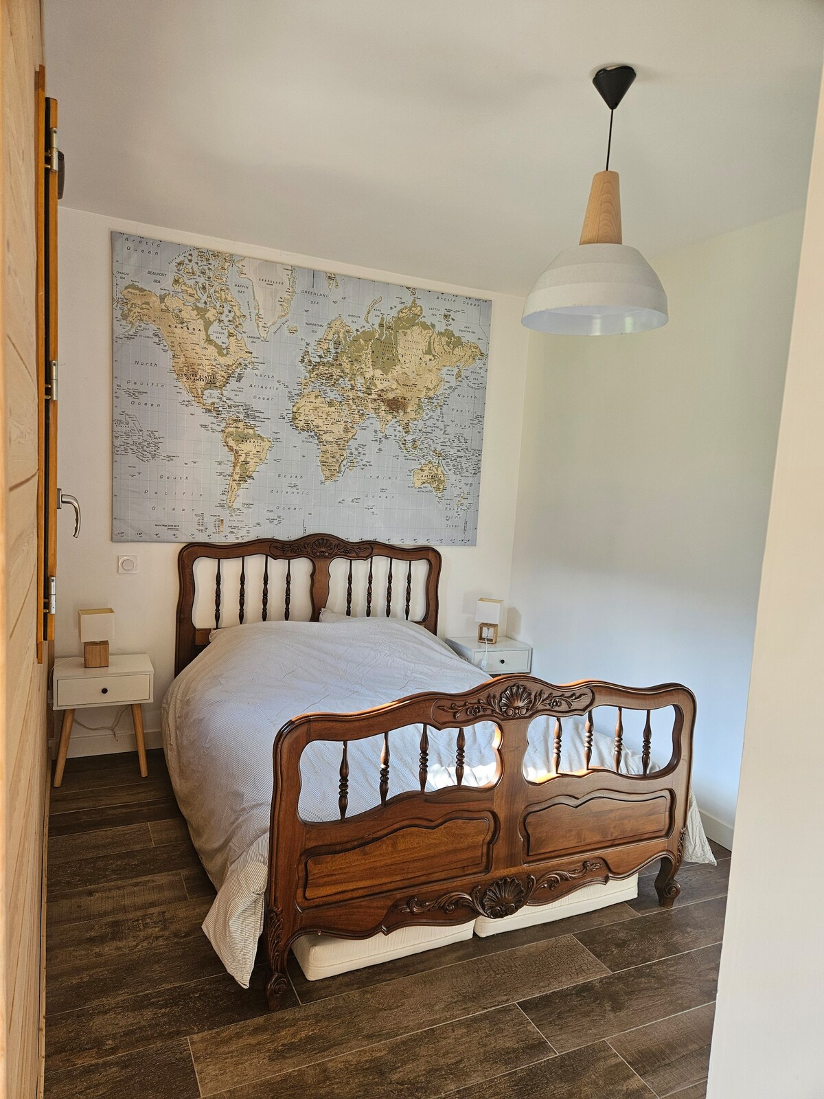 A private bedroom features a traditional wooden bed adorned with a neutral-toned bedding set. Nightstands flank the bed, each with a lamp. A world map decorates the wall above, providing a subtle focal point. The room is illuminated by natural light.