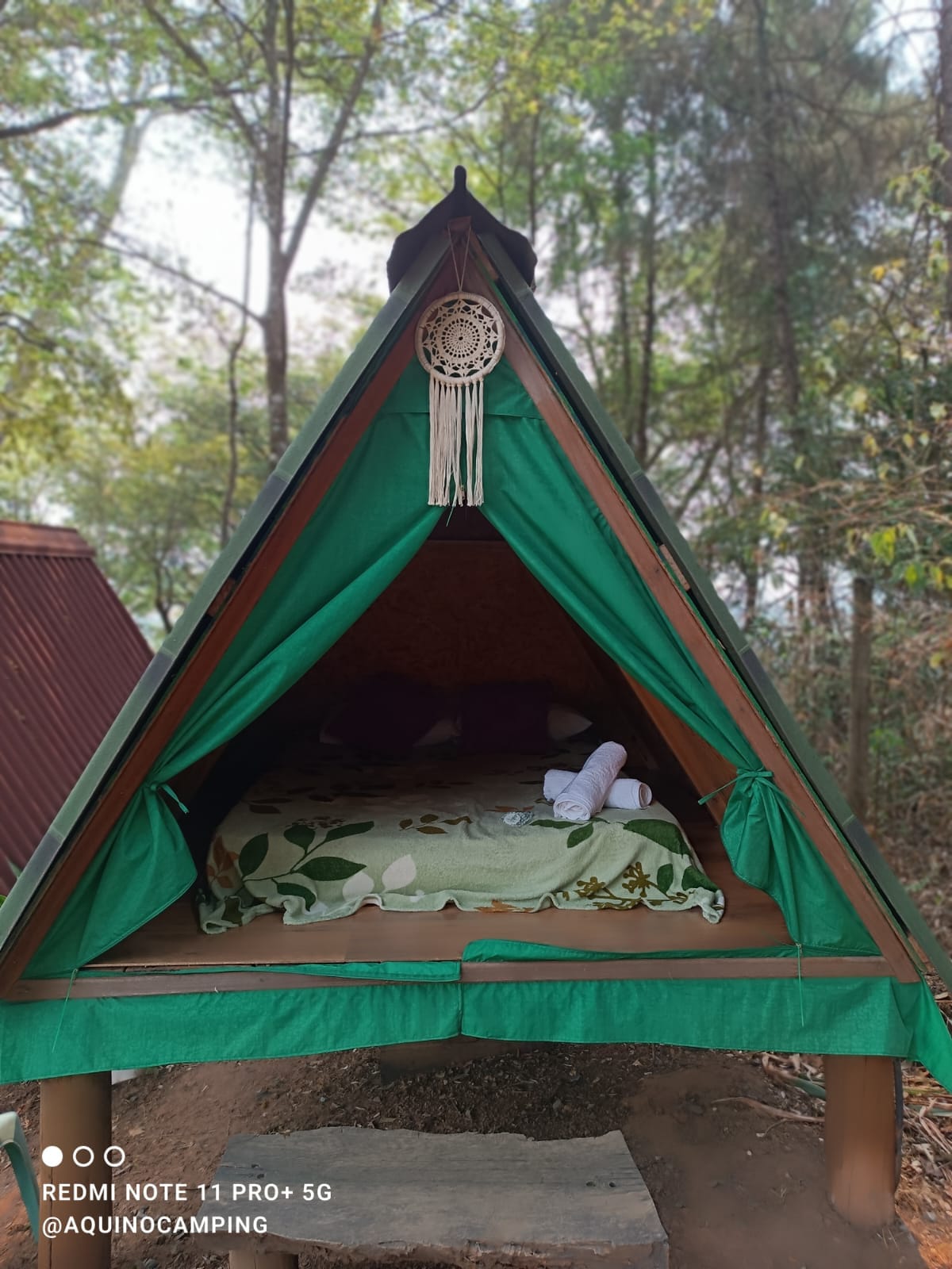 A charming wooden cabin features a steeply pitched roof and is surrounded by greenery. The inviting sleeping area is adorned with a floral-patterned blanket and neatly arranged towels, offering a connection to nature in a cozy setting.