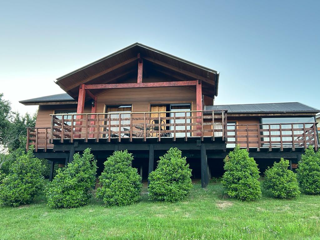 A charming house is set on raised wooden supports, featuring a spacious balcony with railings. The exterior is composed of warm-toned wood, complemented by neatly trimmed shrubs in the front yard. The roof has a distinctive overhang, offering architectural interest.