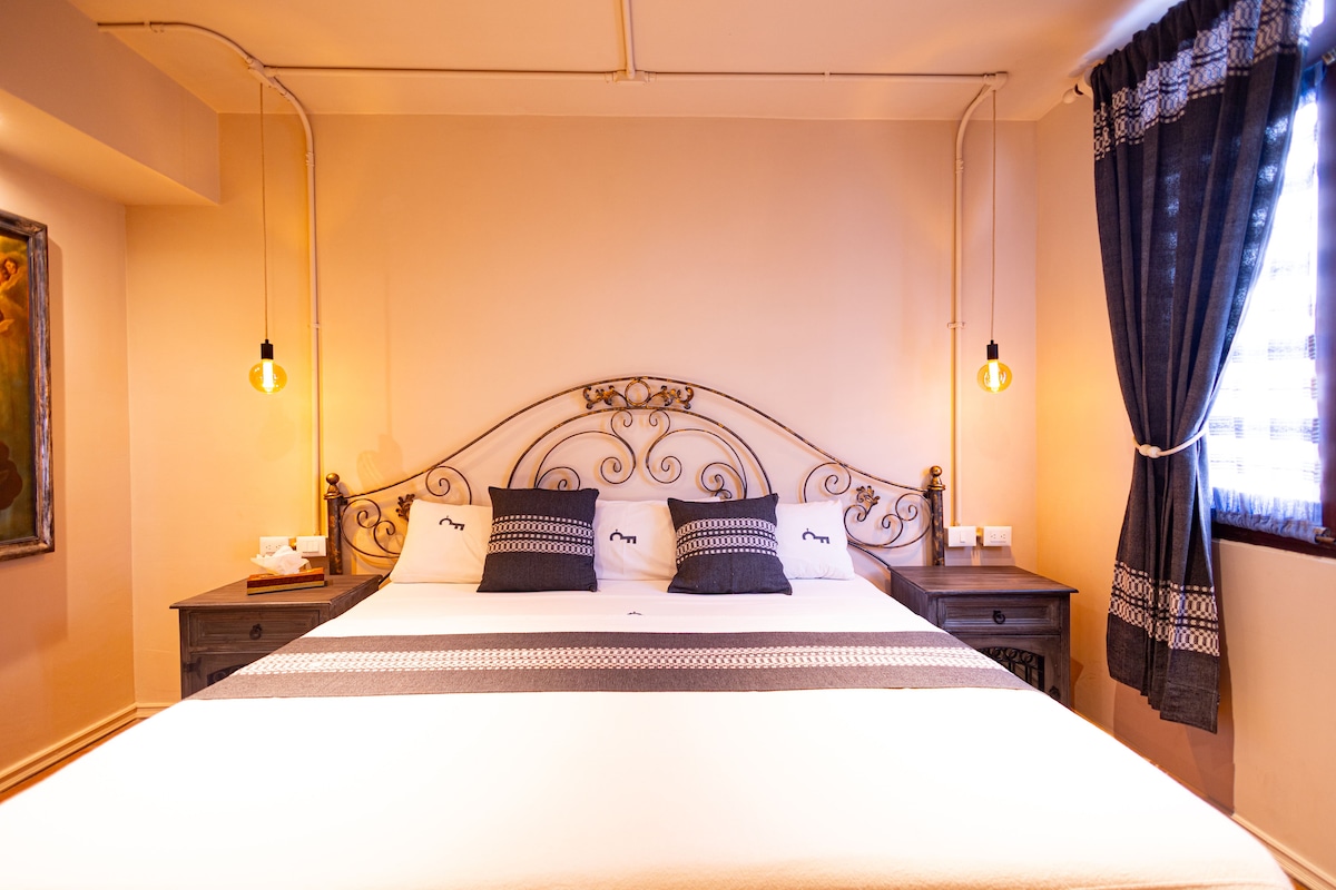 A spacious bed is positioned against a decorative metal headboard, complemented by two bedside lamps. Dark curtains frame a partially open window, allowing soft natural light to fill the room. Neutral-colored walls create a calm environment, enhancing the overall comfort.