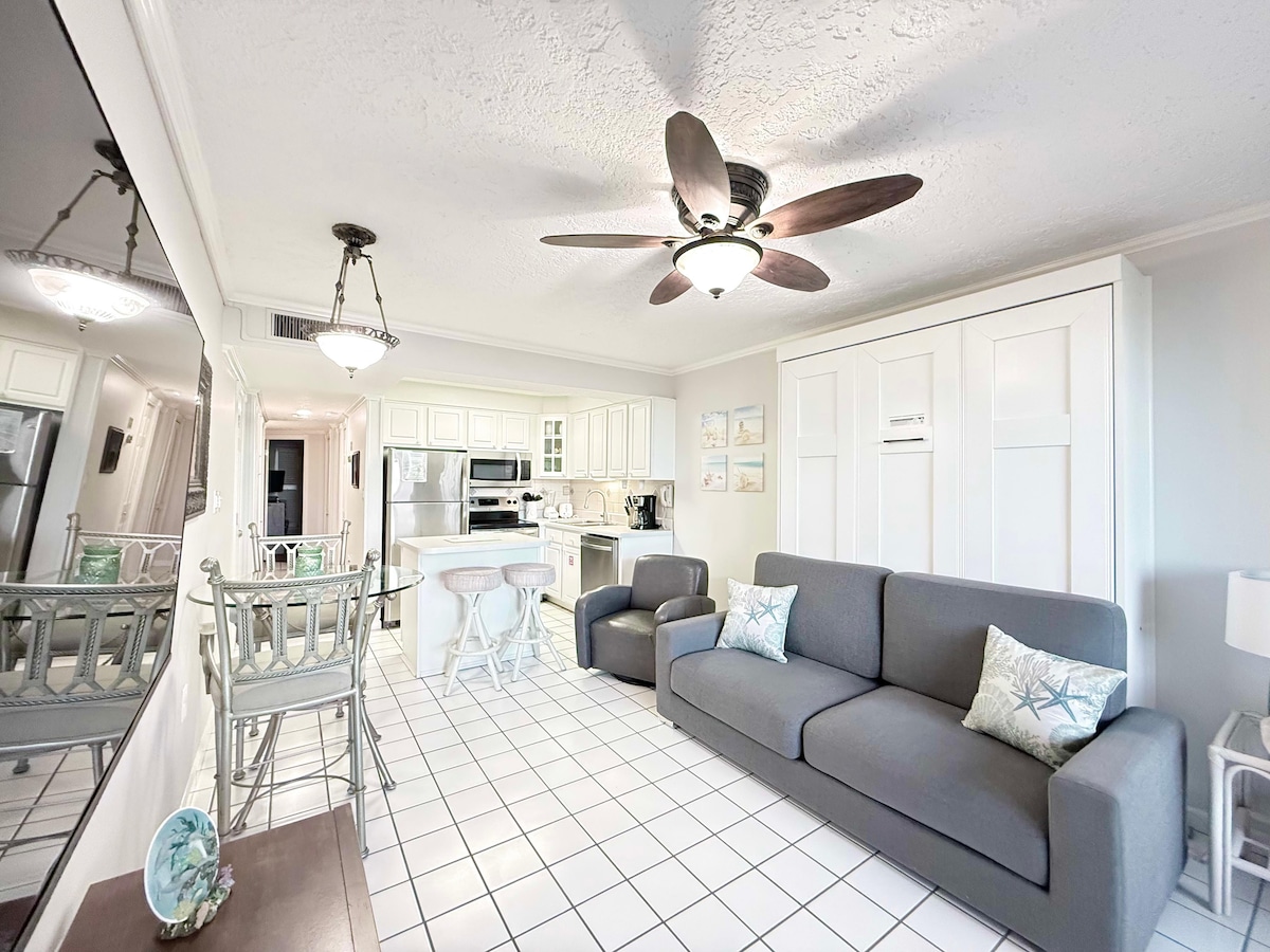 A spacious living area is highlighted by a comfortable grey sofa, matching chairs, and a large mirror that reflects the open layout. The dining area includes a light wood table with soft blue accents. A ceiling fan is centered above, complementing the bright, tiled flooring.