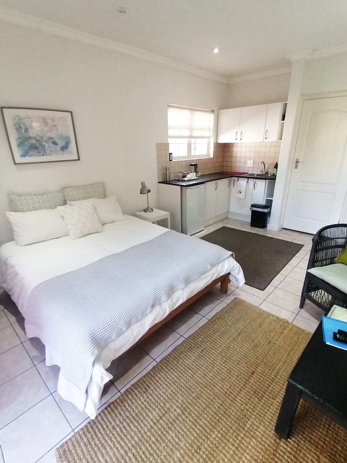 A cozy studio flat features a spacious bed with neutral bedding and several decorative pillows. A kitchenette is visibly equipped with essential appliances. Natural light enters through a window, illuminating the area, which is complemented by a small rug and a coffee table.