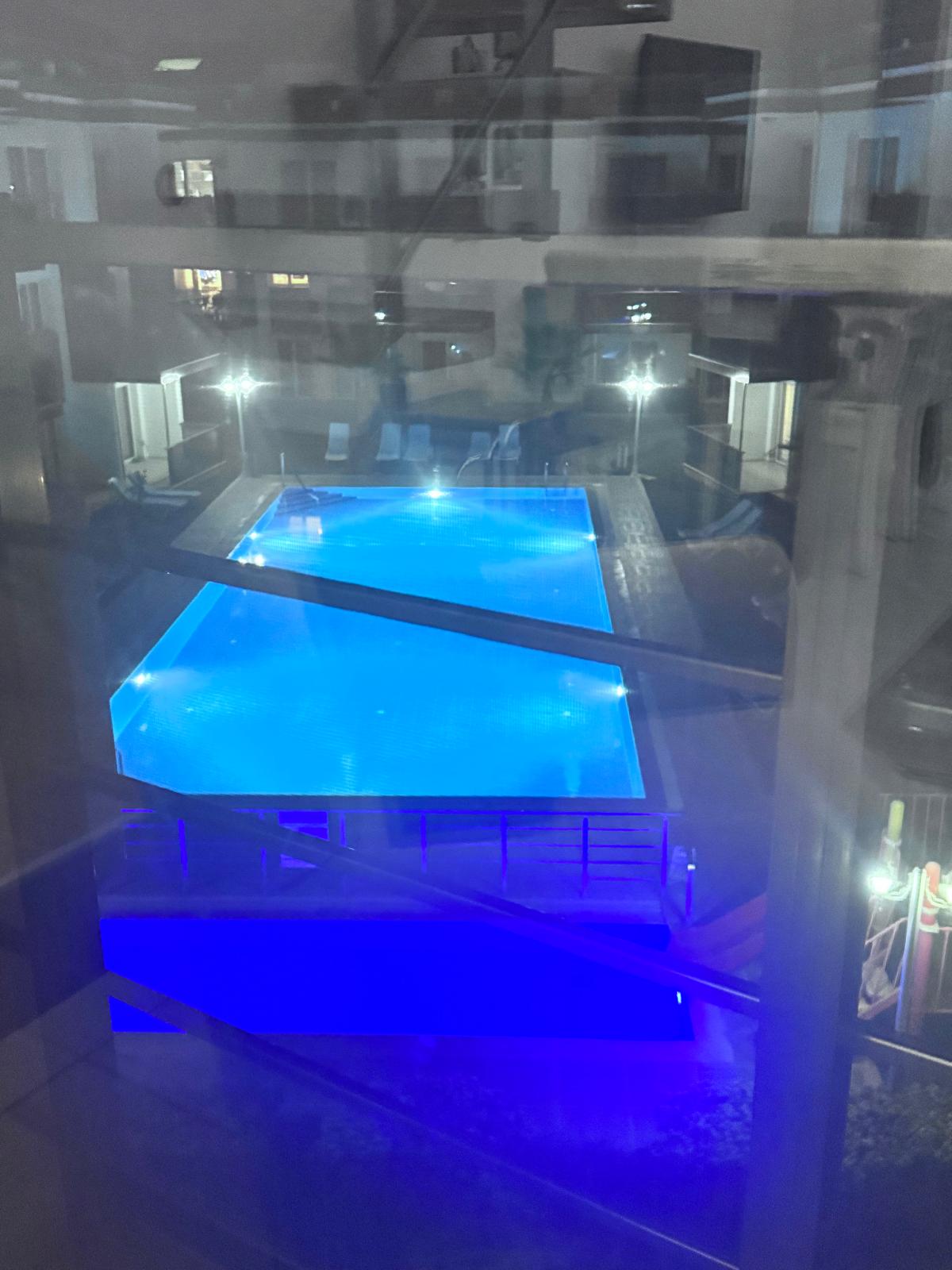 A spacious swimming pool is illuminated in blue light, surrounded by lounge chairs and wooden decking. The scene is set at night, with soft lighting accentuating the tranquil environment. Views are captured through a window, showing the pool's inviting atmosphere.