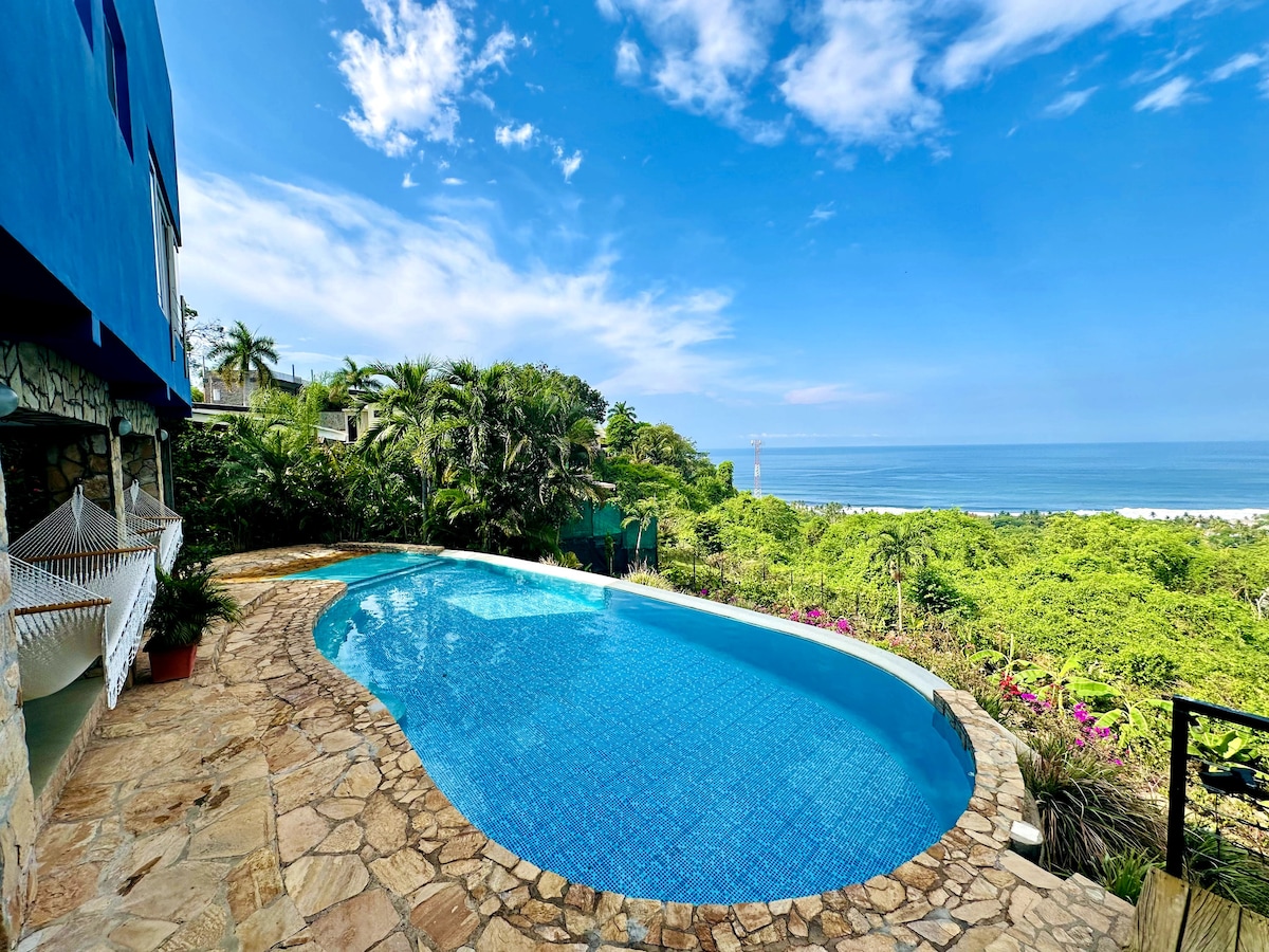 An ocean-facing infinity pool showcases clear blue waters, set against a backdrop of lush greenery and the ocean. The surrounding stone patio features natural stone tiles, enhancing the outdoor space. A hammock is visible, inviting relaxation by the pool's edge.