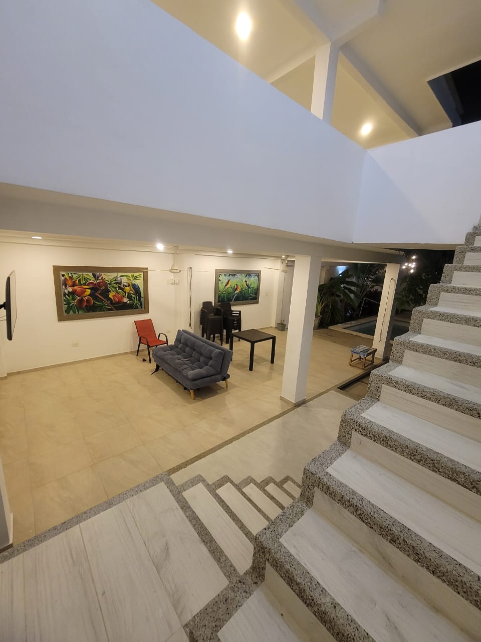 A spacious living area is visible, featuring modern furnishings including a gray sofa and a black dining table. Colorful artwork adorns the walls, enhancing the atmosphere. Natural light filters in from overhead, illuminating the room's clean lines and open layout, which connects to a staircase.