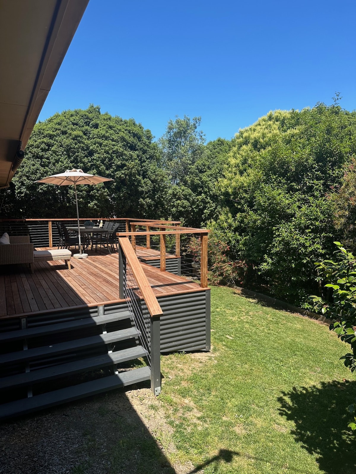 A spacious outdoor deck is shown, surrounded by lush greenery. A large table and chairs are positioned under a parasol, while a lounge area is visible, offering a place to relax and enjoy the tranquil garden views.