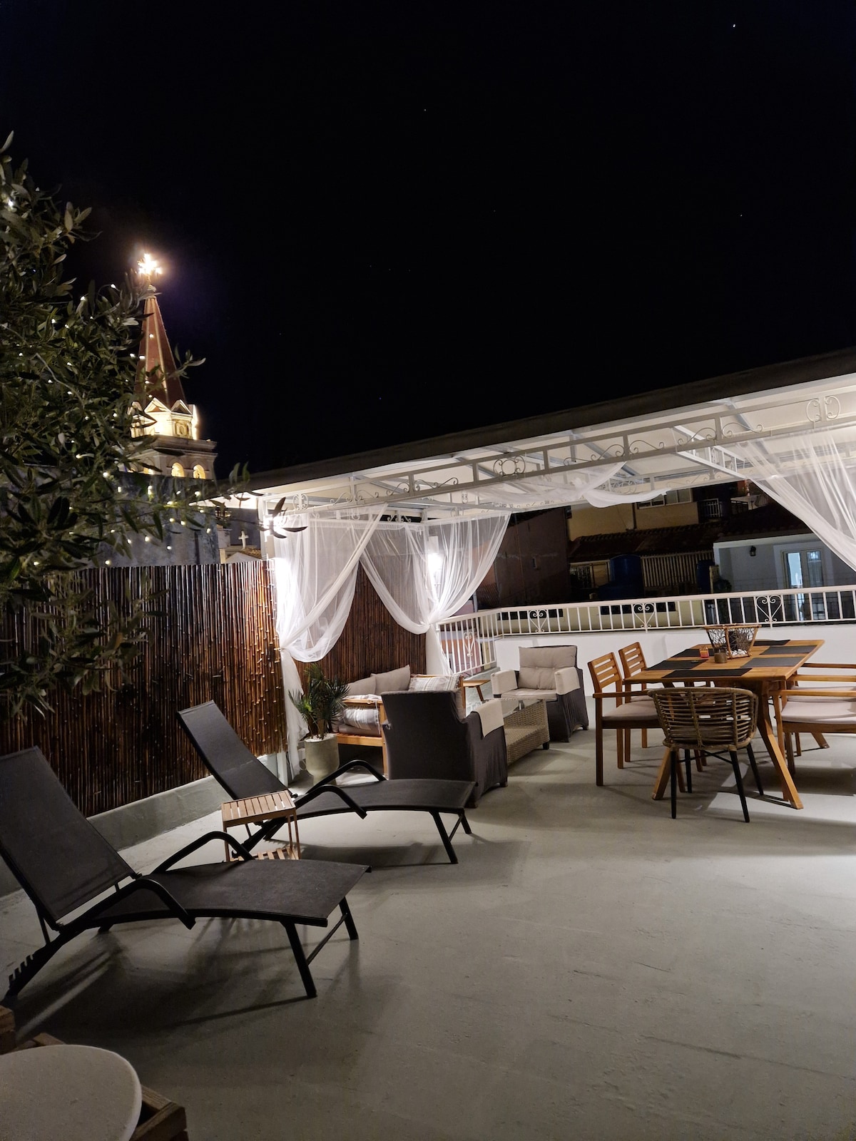 A rooftop terrace is illuminated at night, featuring a dining table surrounded by wooden chairs. Two loungers are positioned to the side, with decorative curtains providing a gentle touch. The background shows a church steeple lit against the night sky, enhancing the serene atmosphere.