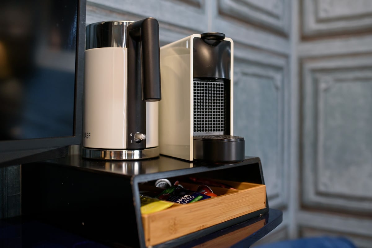 An electric kettle and a coffee maker are positioned on a sleek countertop. A wooden drawer reveals an assortment of tea bags and coffee pods. The backdrop features textured metallic panels that add subtle character to the setting.