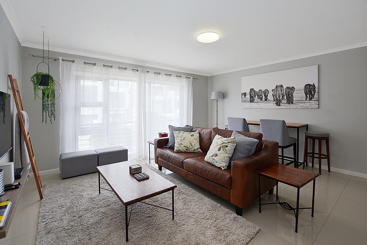 A comfortable living area is presented with a brown leather sofa and two light gray chairs. A wooden coffee table and side tables complement the space. Large windows allow natural light to brighten the room, which features neutral wall colors and a decorative artwork depicting elephants.