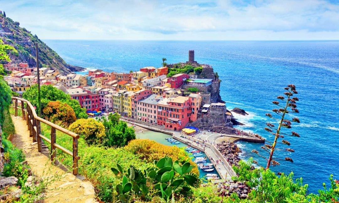A coastal view showcases a colorful village overlooking the sea, with vibrant buildings nestled against the rocky shoreline. The tranquil blue water gently laps at the shore, while green hills flank the scene. A path leads down towards the harbor, framed by lush vegetation.
