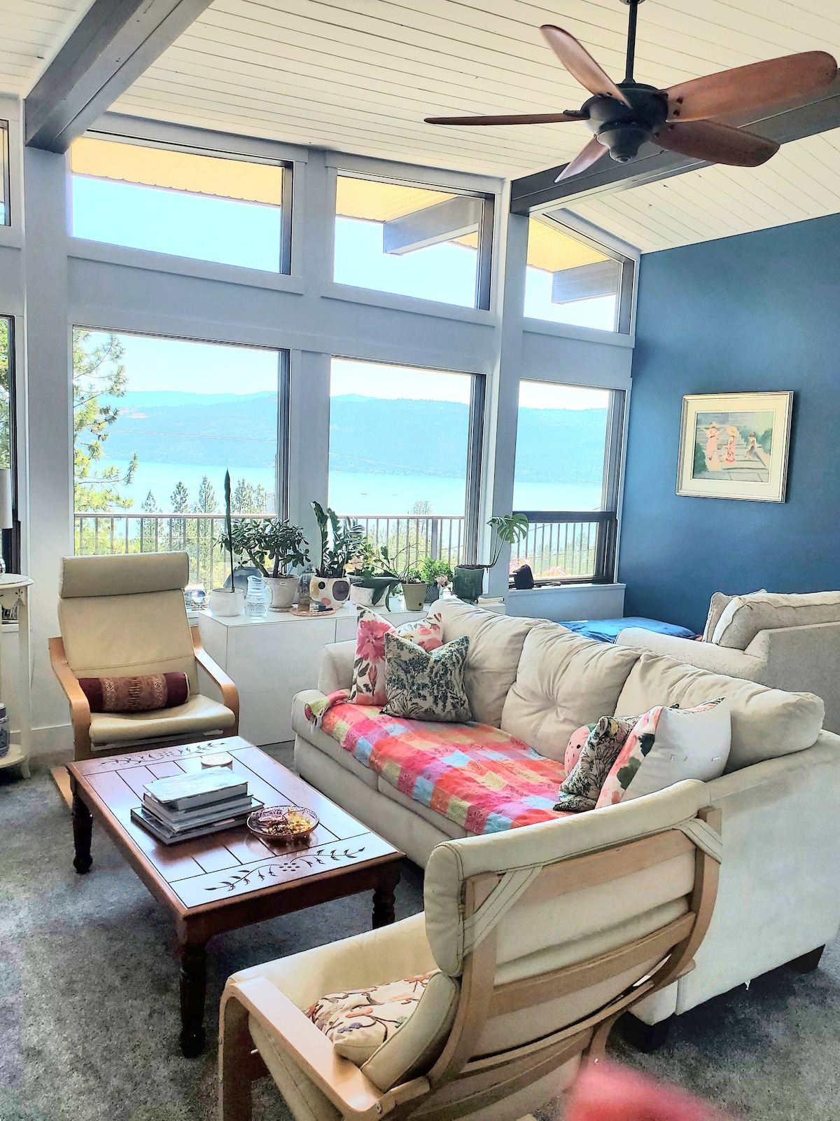 A bright living area showcases large windows offering views of Lake Okanagan. A beige sofa, adorned with colorful throw pillows and a multi-colored blanket, faces a wooden coffee table. A light-toned chair and various plants enhance the inviting atmosphere.