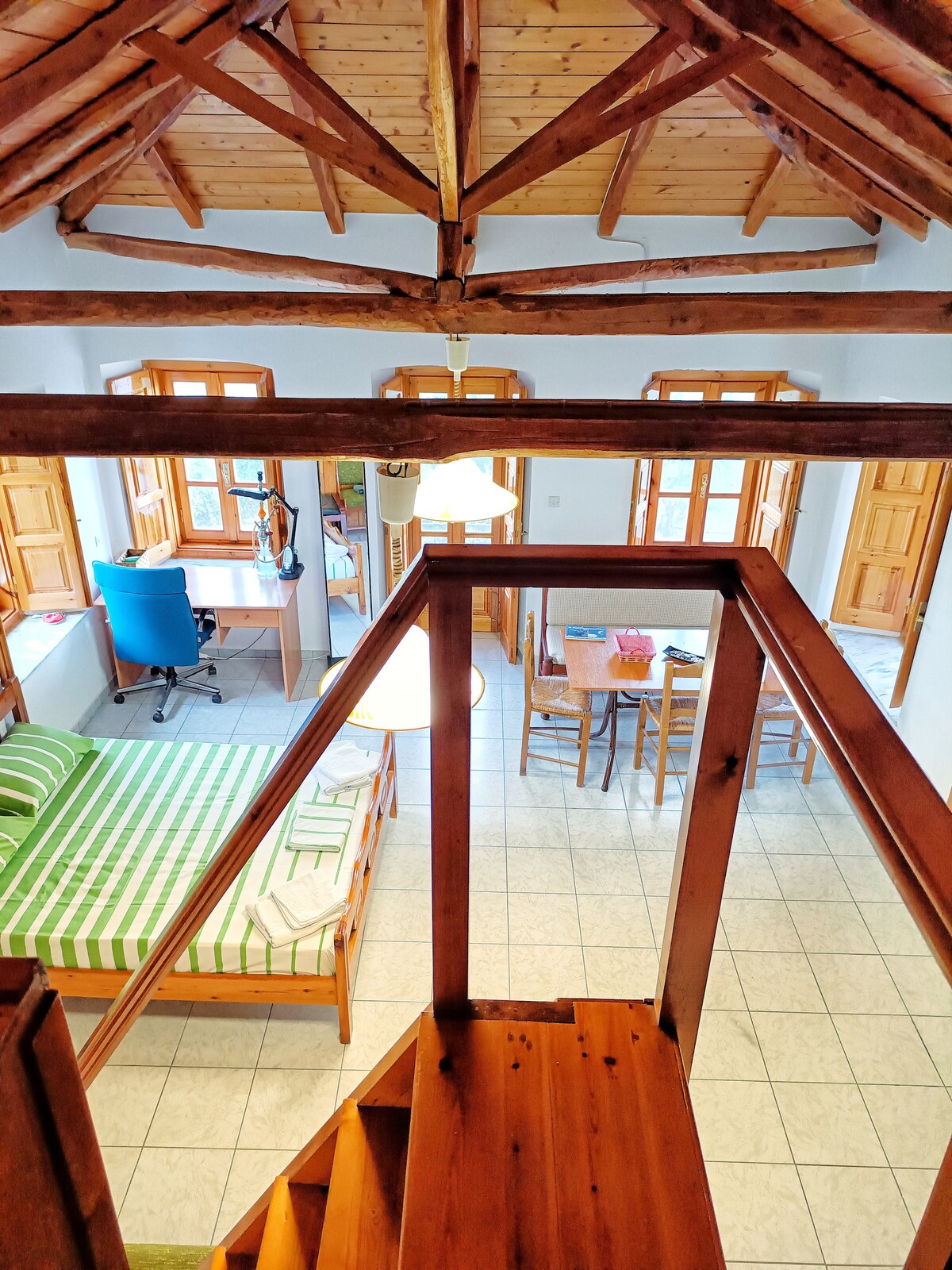 A spacious loft area affords a view of the open floor plan below, featuring a large bed with green and white striped bedding. Natural light enters through multiple windows, highlighting the wooden beams and the dining area with a table and chairs.