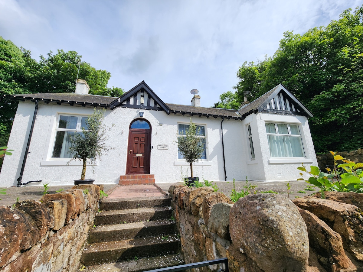 The exterior of a detached cottage is showcased, featuring white walls and black trim. A welcoming front door is flanked by small olive trees. Steps lead to the entrance, with a stone wall providing a natural boundary and lush greenery surrounding the property.