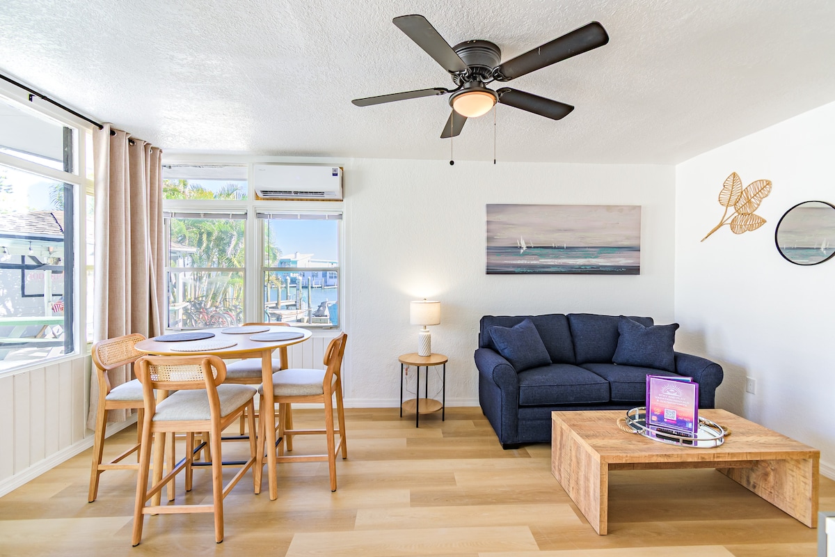 A living area features a comfortable blue sofa and a wooden coffee table. A round dining table with four chairs is positioned nearby. Large windows offer natural light and a view of the outdoors, while a ceiling fan adds to the room's functionality.