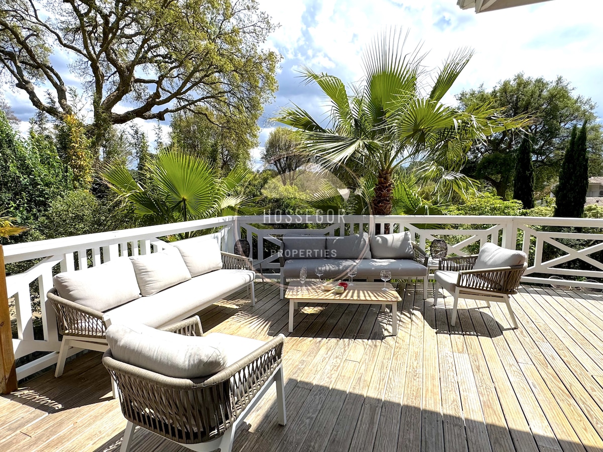 An outdoor seating area on a wooden deck is presented, featuring a comfortable sofa set and low table surrounded by lush greenery. Tall palm trees add a tropical touch, creating an inviting space for relaxation in the fresh air.