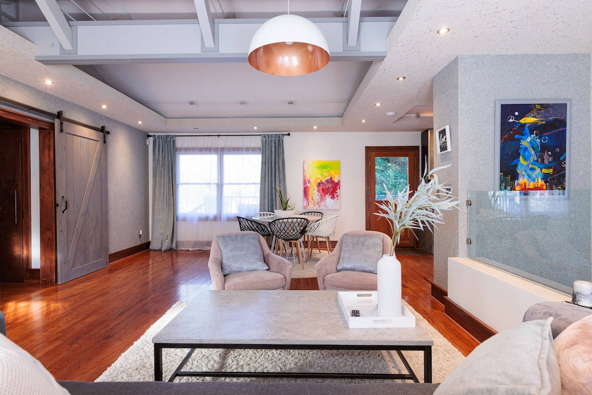 An open-concept living area is illuminated by soft lighting fixtures. Two comfortable armchairs sit across from a modern coffee table. A dining area with sleek chairs is visible in the background, complemented by a colorful wall art piece and large windows adorned with curtains.