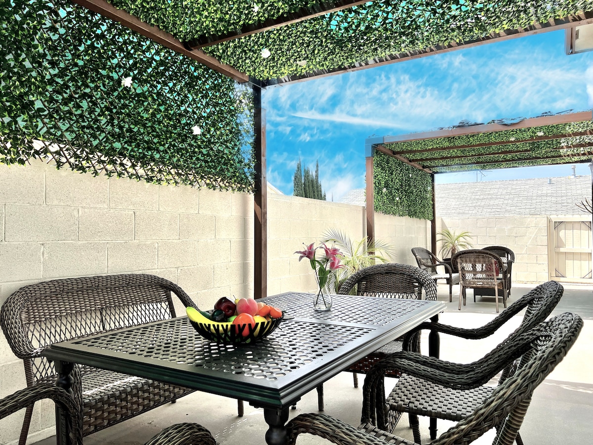 A shaded patio area showcases a large dining table surrounded by woven chairs. A bowl of fresh fruit is centered on the table, while potted plants add greenery to the space. The surrounding walls are adorned with climbing plants, enhancing the natural ambiance.