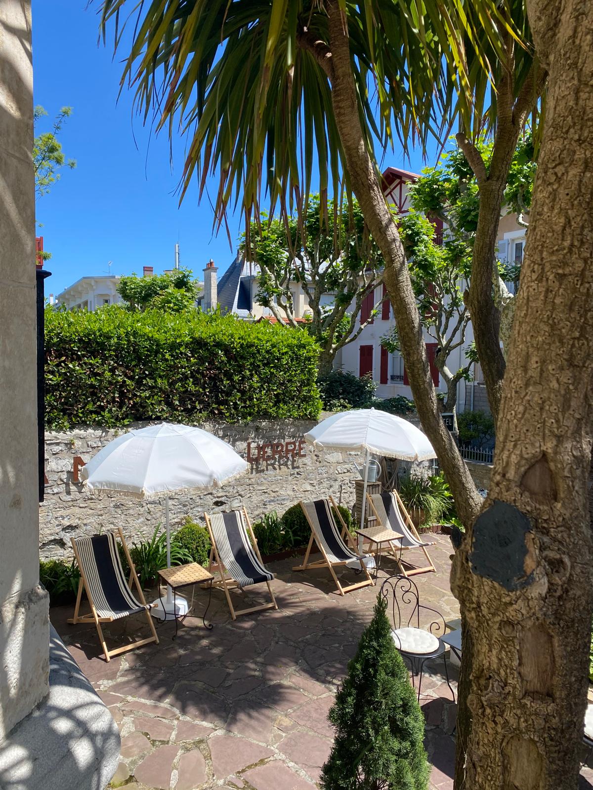 A serene outdoor area is presented, featuring several deck chairs arranged under white umbrellas. Lush greenery surrounds the space, with a stone pathway leading to the seating area. The vibrant blue sky adds a bright backdrop to the peaceful garden setting.