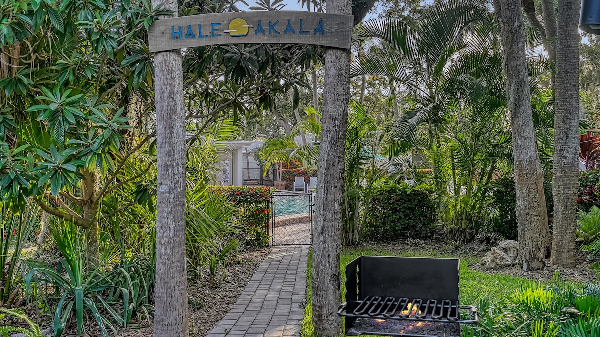 An entrance sign reading 'Hale Akala' is framed by lush tropical foliage. A path of stone pavers leads to a community pool visible through a gated entry. To the side, a grill is positioned in a designated area surrounded by greenery.