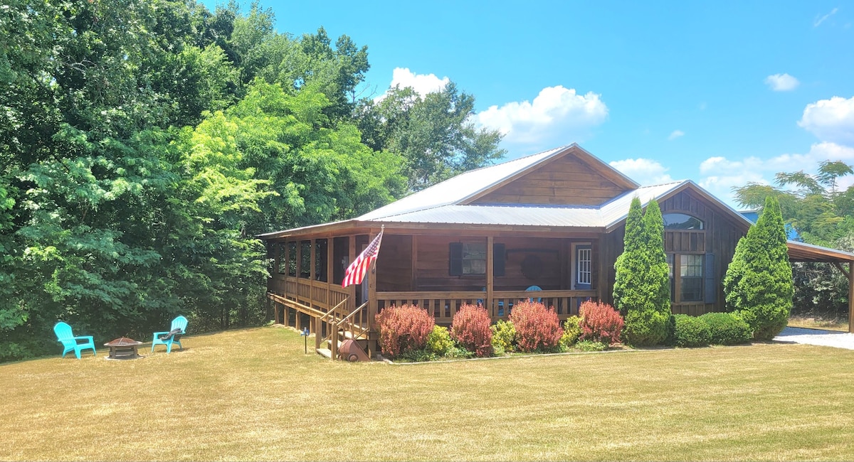 A rustic cabin is set against a backdrop of lush greenery. A large deck extends across the front, complemented by vibrant landscaping. Two teal chairs are situated near a fire pit in the grassy yard, while an American flag is displayed on the porch.