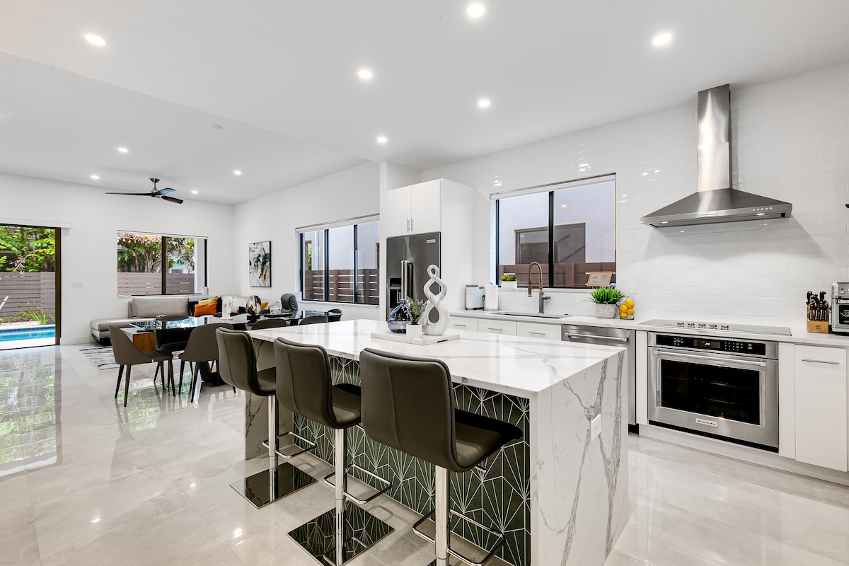 A modern kitchen is presented with marble countertops and sleek cabinetry. Bar stools line the island, complemented by large windows that invite natural light. Stainless steel appliances and a stylish range hood are visible, offering a contemporary cooking space. The open floor plan connects to the outdoor area.