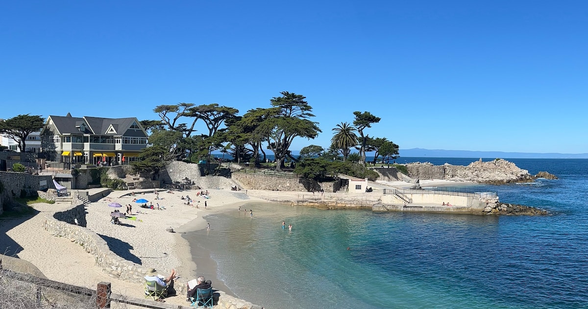 A serene beach scene is presented, featuring soft sandy shores and calm turquoise waters. The area is dotted with sunbathers and beach-goers. Lush trees provide shade, and cozy seating is visible in the foreground. Nearby buildings and rocky formations enhance the coastal view.