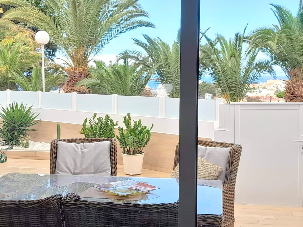 An outdoor terrace features a dining table surrounded by comfortable seating. Lush greenery, including palm trees and potted plants, provides a scenic backdrop, with glimpses of the sea visible beyond the fencing. Natural light enhances the inviting atmosphere.