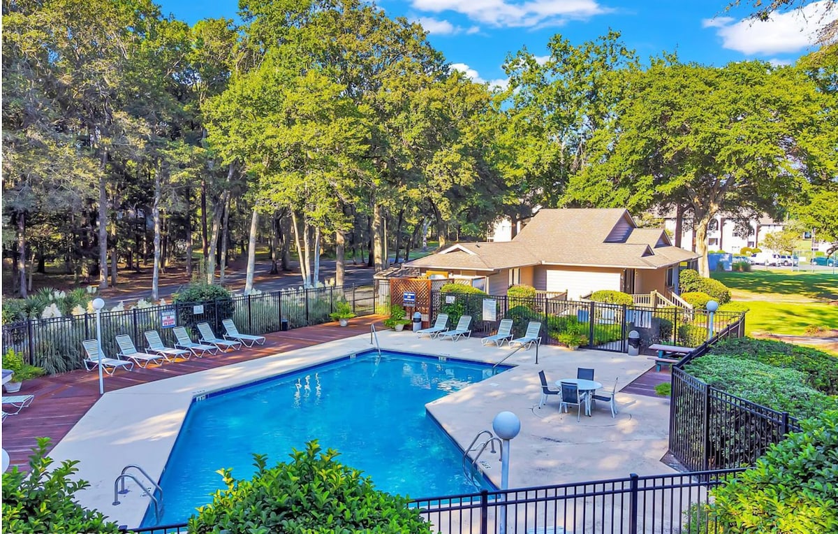The pool area is surrounded by lush greenery and tall trees, offering a serene retreat. Loungers frame the large pool, with a shaded seating area nearby. A small building serves as a changing area. Clear blue water reflects the sky, enhancing the inviting atmosphere.