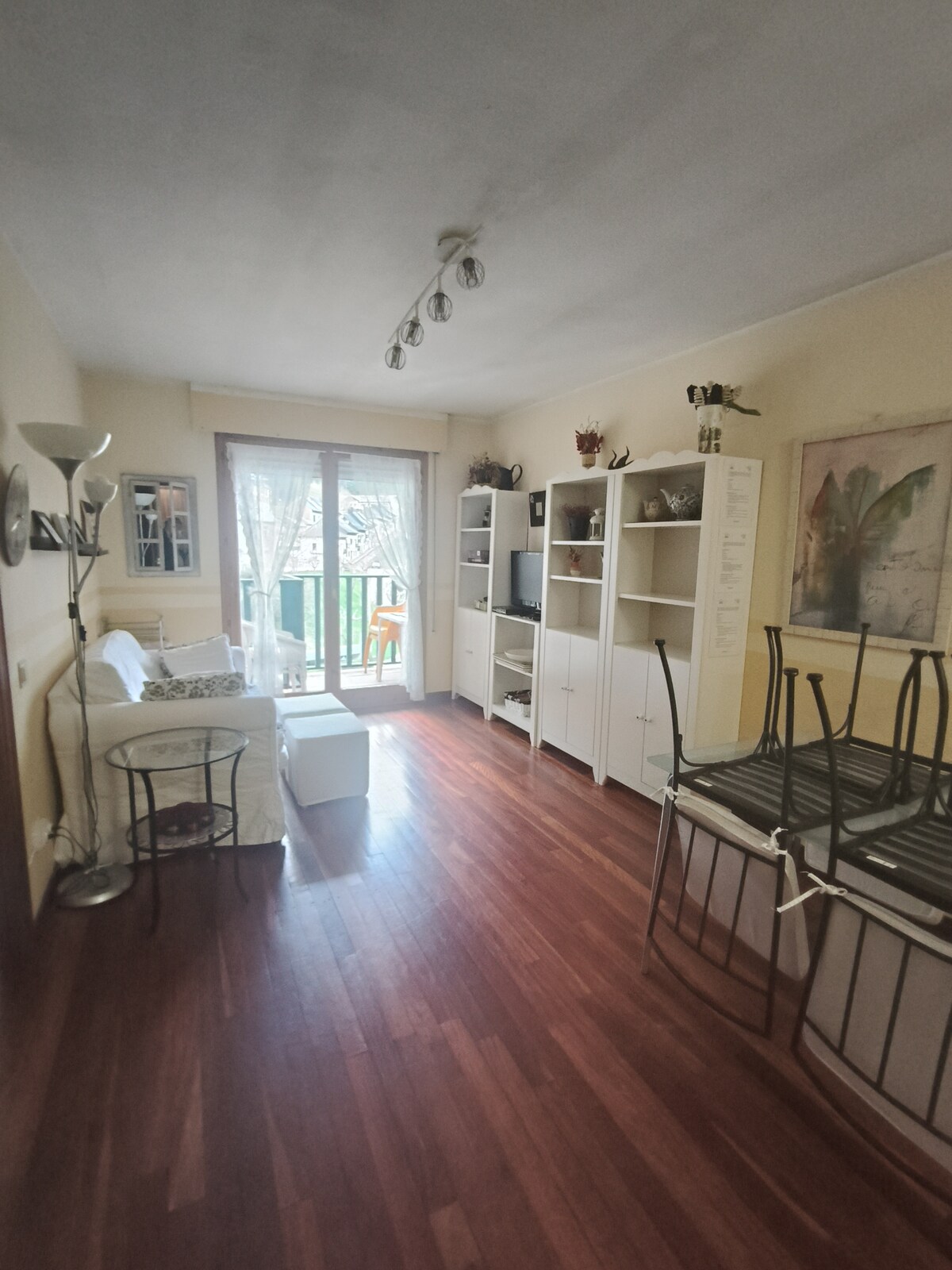 An inviting living area is showcased, featuring a light-colored sofa and a small side table. Storage shelves line one wall, displaying decorative items. Double glass doors lead to a small balcony, allowing natural light to enhance the warm wooden floors.