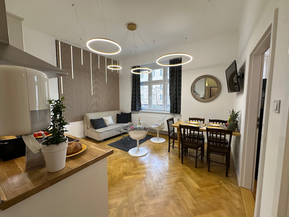 The living area features a light wooden floor and a stylish sofa with round coffee tables. Four pendant lights hang above, illuminating the space. A dining table with four chairs is seen next to a mirror, while a flat-screen TV is mounted on the wall.