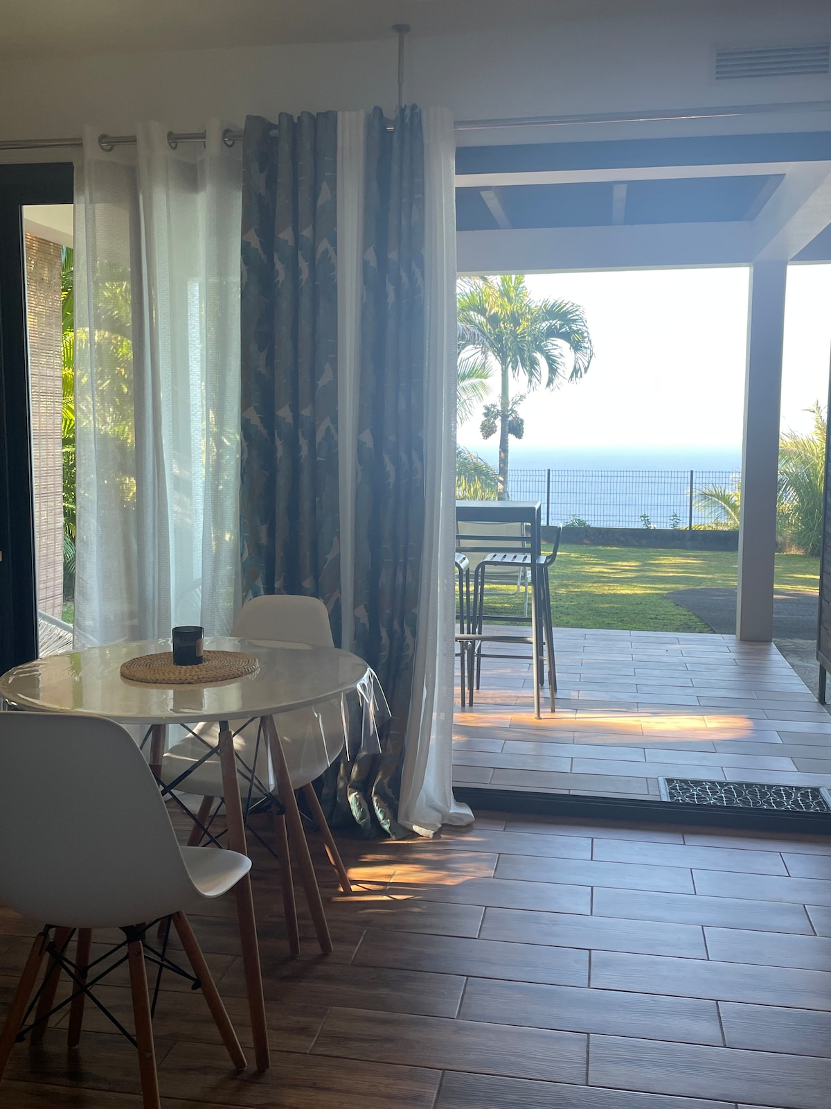A dining area features a round table with white chairs, positioned near sliding glass doors that open to a terrace. Outside, a verdant view includes palm trees and ocean scenery. Natural light fills the space through sheer curtains, enhancing the inviting atmosphere.