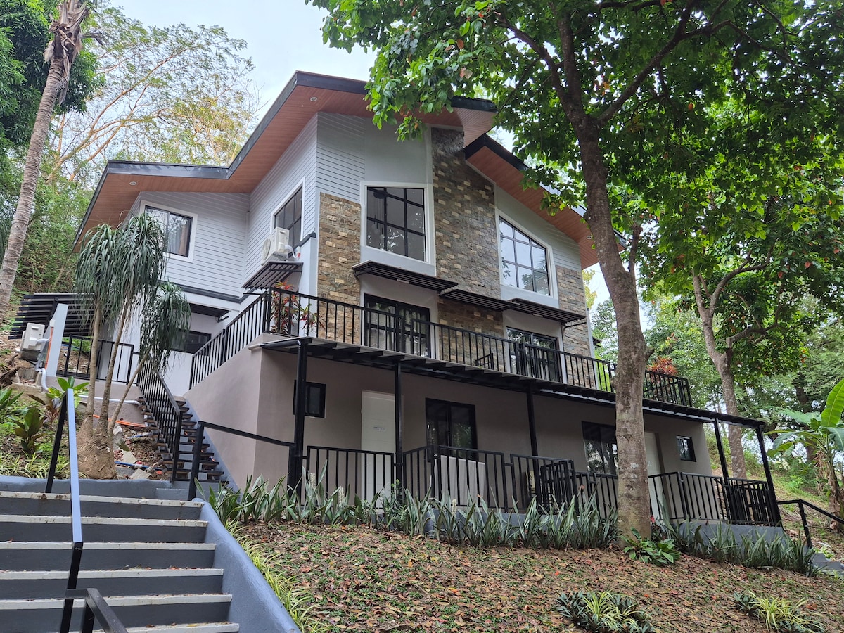 A modern two-story house is nestled among lush greenery, featuring a blend of stone and wood in its exterior design. A balcony extends across the front, enhancing outdoor enjoyment. Steps lead to the entrance, surrounded by well-maintained landscaping and tall trees.
