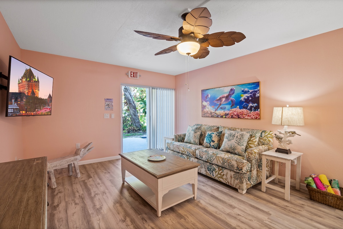 A living room features a comfortable sofa with a floral pattern and a light wood coffee table. A flat-screen TV is mounted on the wall. Natural light streams through sliding glass doors, leading to an outdoor space. A decorative ceiling fan adds an airy feel to the room.