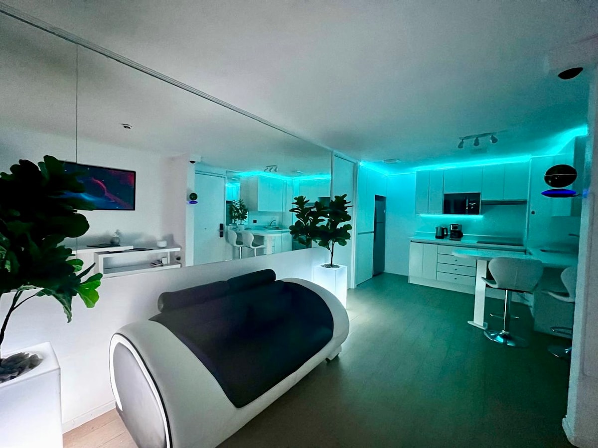 A modern living space features sleek, white furnishings and a mirrored wall that adds depth. Soft blue ambient lighting outlines the kitchen and bar area, while lush green plants offer a touch of nature. The inviting sofa provides a comfortable focal point.