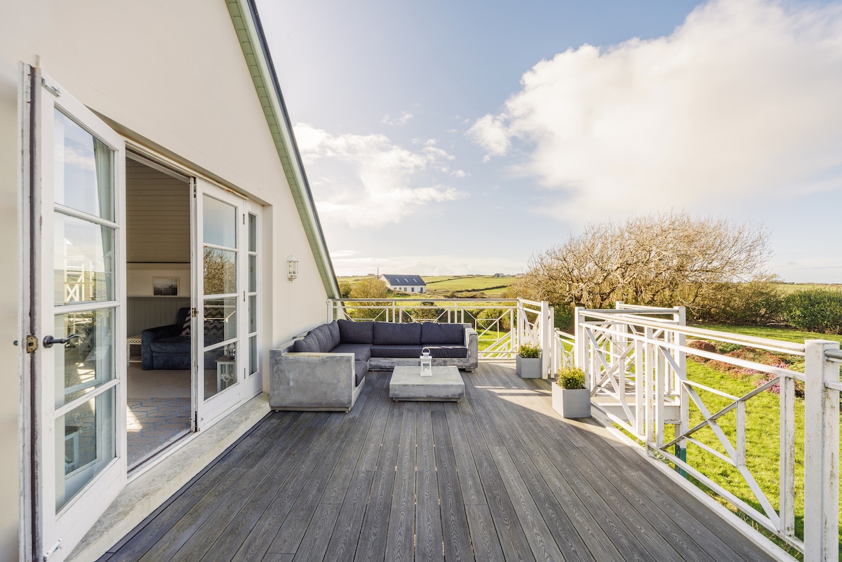 A spacious balcony features elegant outdoor seating, including a large couch and a coffee table. The floor is made of wooden planks, and plants are positioned around the area. A scenic view of the surrounding landscape and sky is visible in the background.